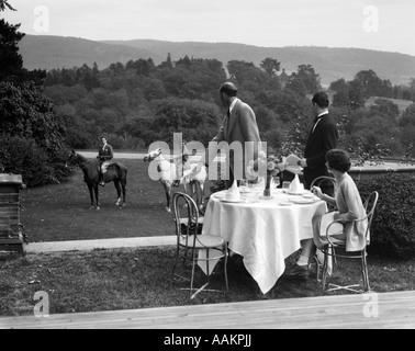 1930s COUNTRY CLUB WITH COUPLE EATING OUTSIDE & ANOTHER COUPLE ON HORSEBACK Stock Photo