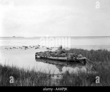 1920s MAN DUCK HUNTER WAITING IN CAMOUFLAGED SNEAKBOX DUCK BOAT BARNEGAT BAY NEW JERSEY USA Stock Photo