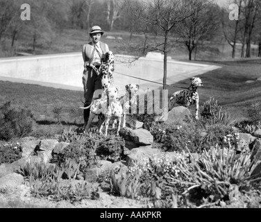 1930s SPORTY WOMAN WEARING JODHPURS WITH 4 DALMATIANS ON A TANDEM LEASH IN A WOODED PARK NEAR A SWIMMING POOL Stock Photo