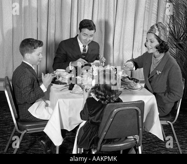 1960s SMILING HAPPY FAMILY OF FOUR EATING MEAL IN RESTAURANT MOM WEARING TURBAN STYLE HAT ADDRESSING HER DAUGHTER Stock Photo