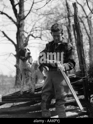 1930s MAN BIRD HUNTER HOLDING DOUBLE BARREL SHOTGUN LOADING SHELLS WITH HIS POINTER DOG STANDING ON A SPLIT RAIL FENCE Stock Photo