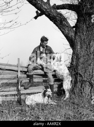1920s 1930s MAN IN JODHPURS & LEATHER JACKET SITTING ON POST & RAIL FENCE UNDER TREE WITH HUNTING DOGS Stock Photo