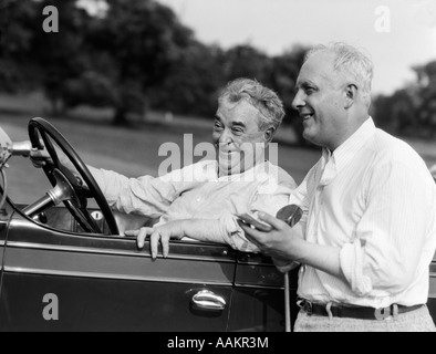 1920s 1930s SENIOR MAN SITTING DRIVING CAR AND ANOTHER STANDING HOLDING GOLF CLUB SHARING A LAUGH Stock Photo