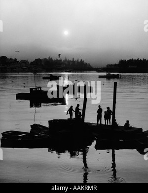 1970s PEOPLE FISHING OFF OF DOCK WITH SMALL BOATS IN BACKGROUND AT DUSK Stock Photo