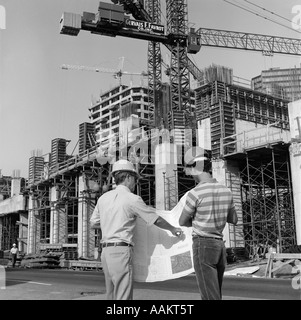1980s REAR VIEW OF TWO HARD HAT WORKERS STANDING IN FRONT OF MAJOR CONSTRUCTION SITE LOOKING OVER BUILDING PLANS MEN OUTDOOR Stock Photo