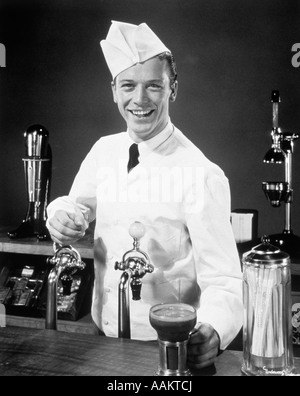 1940s 1950s SMILING YOUNG MAN SODA JERK AT COUNTER SERVING AN ICE CREAM SODA LOOKING AT CAMERA