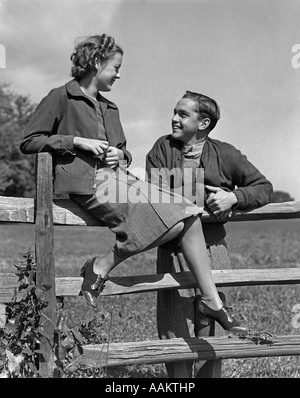 1930s 1940s YOUNG SMILING TEENAGE COUPLE TALKING GIRL SITTING ON WOODEN SPLIT RAIL FENCE Stock Photo