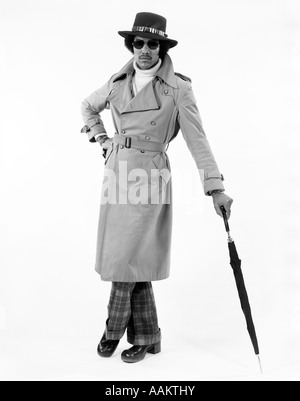 1970s PORTRAIT OF AFRICAN AMERICAN MAN DRESSED IN TRENCH COAT AND HAT LEANING ON UMBRELLA LOOKING AT CAMERA Stock Photo