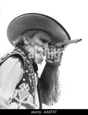 SENIOR BEARDED COWBOY POINTING A REVOLVER AT THE CAMERA DRESSED IN FRINGED GLOVE BEADED VEST WIDE BRIMMED HAT Stock Photo