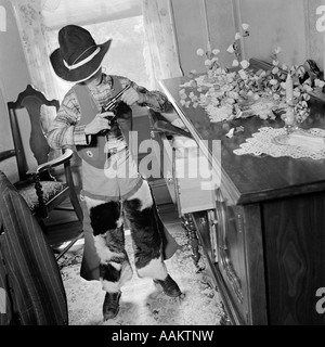 1950s BOY STANDING IN FRONT OF BUREAU DRAWER LOADING TOY GUN DRESSED IN COWBOY CLOTHES COSTUME Stock Photo