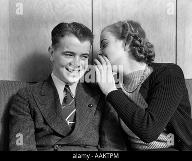 1930s 1940s YOUNG TEENAGE COUPLE SITTING ON COUCH GIRL WHISPERING SECRET INTO SMILING BOY'S EAR Stock Photo