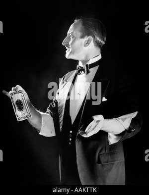 1920s MAGICIAN IN TUXEDO WITH SLEEVES ROLLED UP HOLDING OUT DOLLAR BILL Stock Photo
