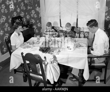 1950s FAMILY OF 5 MOM DAD 3 KIDS SITTING AT DINING ROOM TABLE SAYING GRACE WITH HEADS BOWED Stock Photo