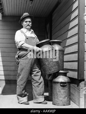 1930s MATURE ELDERLY MAN DAIRY FARMER HOLDING CARRYING METAL MILK CANS CANISTERS FARM LOOKING AT CAMERA Stock Photo
