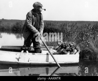 1920s 1930s SENIOR MAN DUCK HUNTER STANDING IN SNEAK-BOX BOAT ABOUT TO DEPLOY DECOYS IN  BARNEGAT BAY WETLANDS MARSH NEW JERSEY Stock Photo