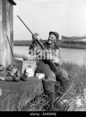 1920s SMILING SENIOR MAN CLEANING PARKER SHOTGUN SURROUNDED BY DUCK DECOYS AMMUNITION BOX BARNEGAT BAY, NEW JERSEY, USA Stock Photo