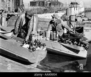1930s 1940s TWO MEN DUCK HUNTERS LOADING SNEAK-BOX BOATS WITH CANADA GOOSE DECOYS AND SHOTGUNS BARNEGAT BAY NEW JERSEY USA Stock Photo