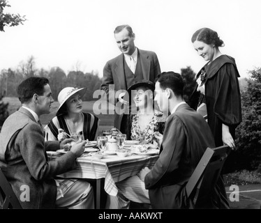 1930s OUTDOOR DINING SCENE TWO UPSCALE COUPLES SEATED AT TABLE ANOTHER COUPLE visiting Stock Photo