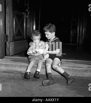 1940s 1950s TWO BOYS SITTING ON FRONT STEPS HOLDING A KITTEN Stock Photo