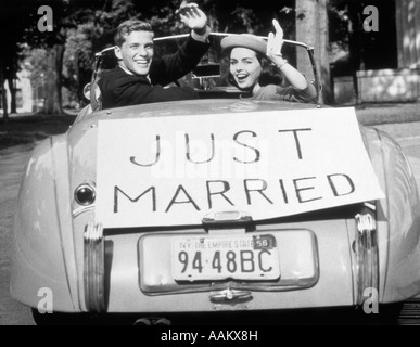 1950s NEWLYWED YOUNG COUPLE MAN WOMAN IN CONVERTIBLE SPORTS CAR WITH JUST MARRIED SIGN WAVING LOOKING AT CAMERA Stock Photo