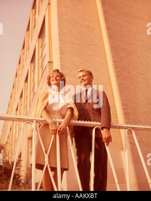 1950s 1960s YOUNG SMILING BLONDE TEENAGE COUPLE STANDING TOGETHER OUTDOORS BY WROUGHT IRON RAILING AND BRICK BUILDING Stock Photo