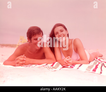 1960s SMILING YOUNG COUPLE LYING SIDE BY SIDE TOGETHER ON BEACH TOWEL Stock Photo