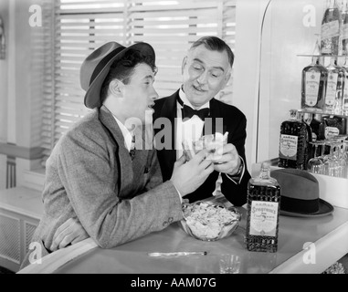1940s 1950s MEN AT BAR DRINKING WHISKEY TOASTING AN UPSCALE GUY IN TUXEDO AND A DRUNKEN BUSINESSMAN Stock Photo
