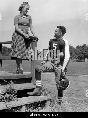 1930s 1940s YOUNG TEENAGE FOOTBALL PLAYER TALKING TO TEENAGE GIRL ON BLEACHER STEPS Stock Photo