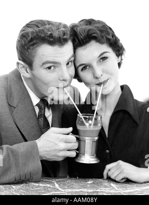1950s SMILING YOUNG COUPLE HEADS TOGETHER SIPPING A SINGLE SODA MILKSHAKE WITH TWO STRAWS LOOKING AT CAMERA