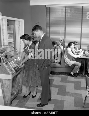 1950s TEEN COUPLE PLAYING JUKE BOX IN MALT SHOP WITH OTHER TEENS IN BOOTHS Stock Photo