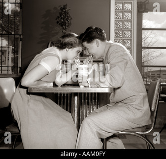 1950s 1960s LAUGHING TEENAGE COUPLE BOY AND GIRL SHARING DRINK TOGETHER WITH TWO STRAWS IN SODA SHOP Stock Photo