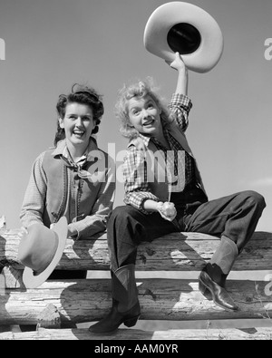 1940s TWO SMILING WOMEN IN COWBOY OUTFITS SITTING ON FENCE ONE RAISING HAT IN AIR OTHER STANDING BESIDE HER BOTH LOOKING AT Stock Photo