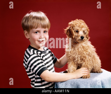 1960s SMILING BLOND BOY STRIPED TEE SHIRT WITH SANDY COLORED DOG PET RED BACKGROUND Stock Photo