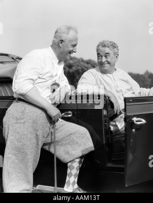 1920s 1930s ELDERLY MAN LEANING ON RUNNING BOARD OF CAR HOLDING GOLF CLUB & TALKING TO ANOTHER MAN SITTING IN THE CAR Stock Photo