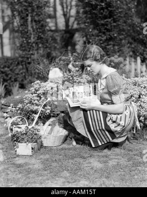 1930s 1940s WOMAN DRESSED IN PRINT DRESS STRIPED APRON KNEELING IN FLOWERS GARDEN READING A GARDENING MANUAL Stock Photo