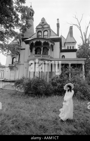 WOMAN IN VICTORIAN COSTUME STANDING ON FRONT LAWN OF LARGE VICTORIAN HOME Stock Photo