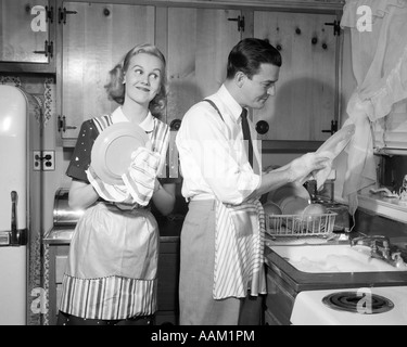 1950s SMILING HAPPY COUPLE MAN AND WOMAN HUSBAND AND WIFE WASHING DRYING DISHES TOGETHER IN KITCHEN Stock Photo