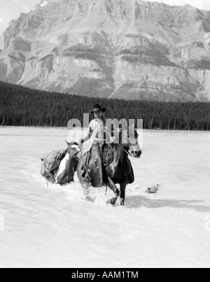 1930s COWBOY WITH BATWING CHAPS ON A BAY HORSE CROSSING A RIVER LEADING A PAINT PACK HORSE WITH MOUNTAINS IN BACKGROUND Stock Photo