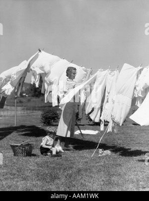 1930s WOMAN IN WHITE COTTON DRESS HANGING A FULL LINE OF CLOTHES IN THE BREEZE WITH A LITTLE BOY & CAT AT HER FEET Stock Photo