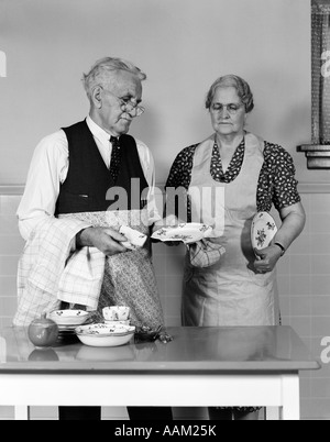 1940s 1950s ELDERLY COUPLE MAN WOMAN IN KITCHEN AT TABLE HOLDING DISHES BOTH WEAR APRON EYE GLASSES Stock Photo