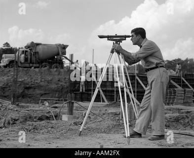 1960s SIDE VIEW OF WORKER SURVEYING CONSTRUCTION SITE WITH CEMENT MIXER IN BACKGROUND Stock Photo