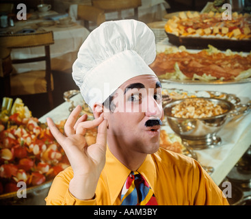 1970s MAN IN CHEF HAT TOQUE MAKE OKAY GESTURE SILLY EXPRESSION FUNNY EYES BROW MOUSTACHE CHARACTER BUFFET BACKDROP Stock Photo