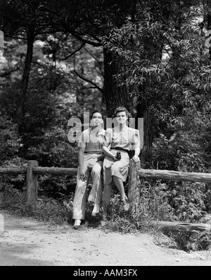 1940s TEEN COUPLE SITTING ON FENCE IN WOODS LOOKING UP FEMALE HOLDING CAMERA Stock Photo