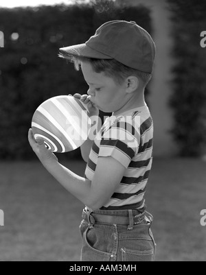 1950s PROFILE OF BOY IN BASEBALL CAP & STRIPED T-SHIRT BLOWING UP STRIPED BALLOON Stock Photo