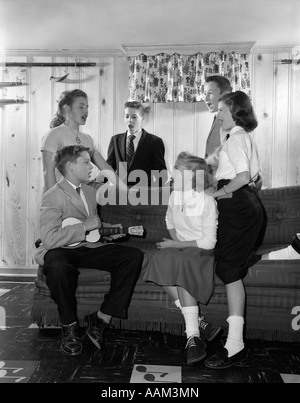 1950s GROUP SIX TEENAGERS THREE BOYS THREE GIRLS SITTING ON COUCH DEN REC ROOM BOY PLAYING UKULELE OTHERS SINGING SONG MUSIC Stock Photo