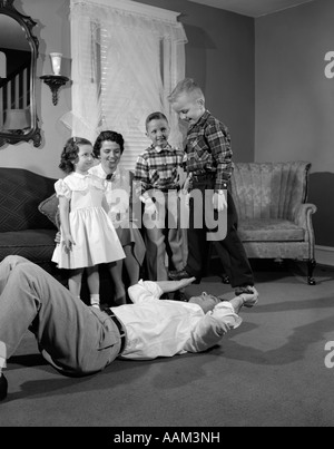 1950s FAMILY FATHER MOTHER DAUGHTER SONS PLAYING GAME FATHER LYING ON BACK LIFTING UP BOY STANDING ON HIS HANDS Stock Photo