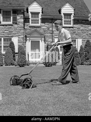 1940s TEEN BOY MOWING GRASS PUSHING MOWER IN FRONT LAWN OF STONE SUBURBAN HOUSE Stock Photo