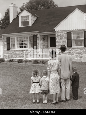 1950s FAMILY OF FIVE WITH BACKS TO CAMERA ON LAWN LOOKING AT FIELDSTONE HOUSE Stock Photo