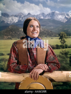 1960s WOMAN IN WESTERN OUTFIT LEANING ON RAIL FENCE HOLDING HAT MOUNTAIN SCENE IN BACKGROUND STUDIO OUTDOOR Stock Photo