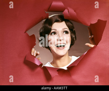 1960s PORTRAIT OF HAPPY YOUNG WOMAN LOOKING THROUGH TORN PAPER JOY SYMBOLIC ONE SMILING Stock Photo